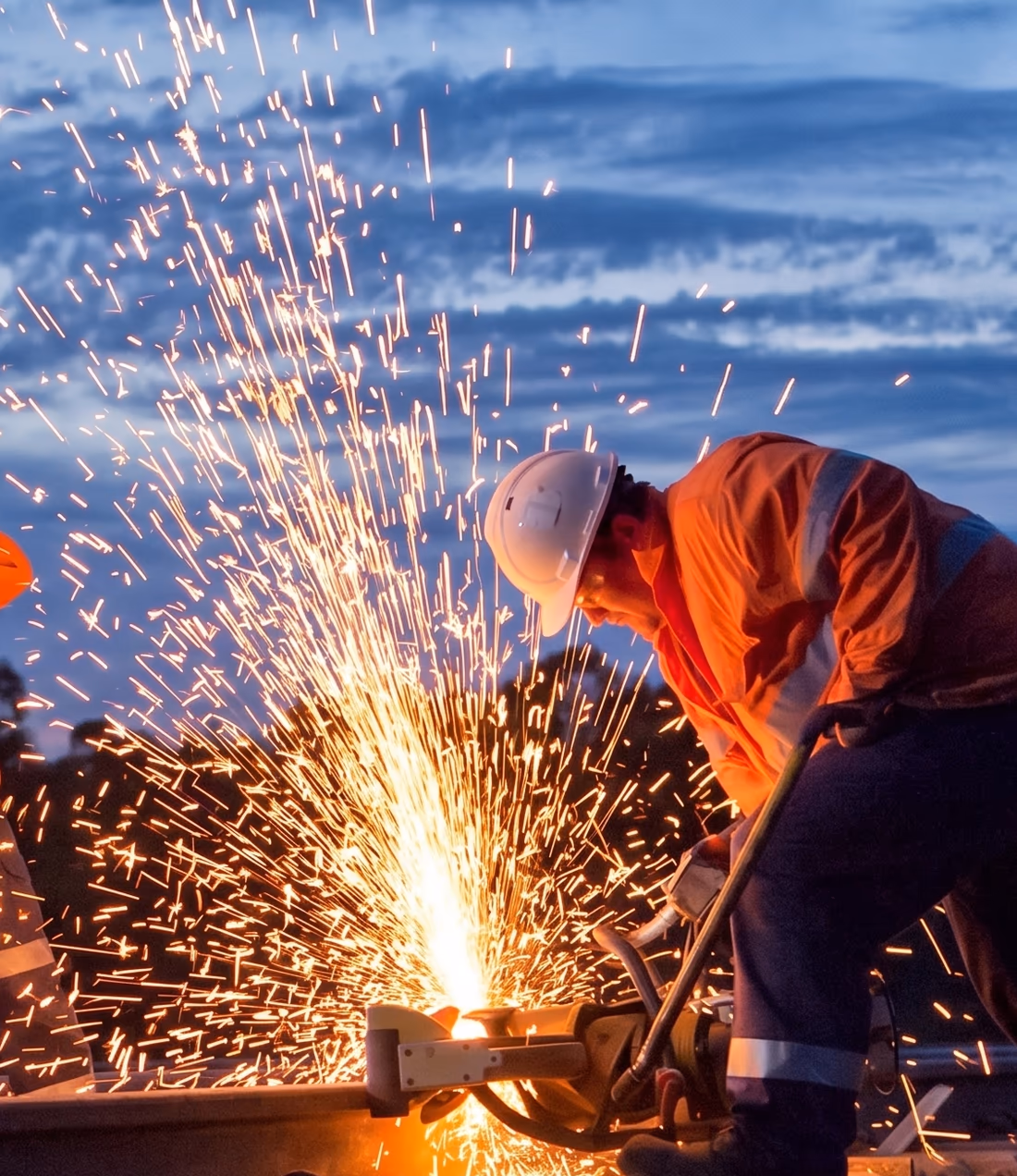 IT support Hunter Valley — a hi-vis boilermaker cutting steel at dusk in a Hunter Valley fabrication yard, sparks arcing from the saw